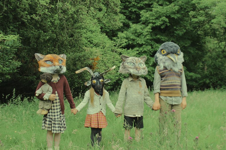 Four children in animal masks standing in field with woods in background